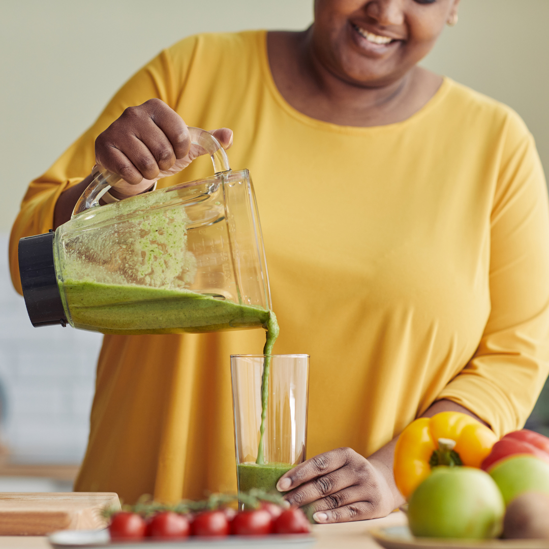 Smiling woman in yellow shirt pouring a green superfood smoothie into a glass with fresh fruits and vegetables on the counter.